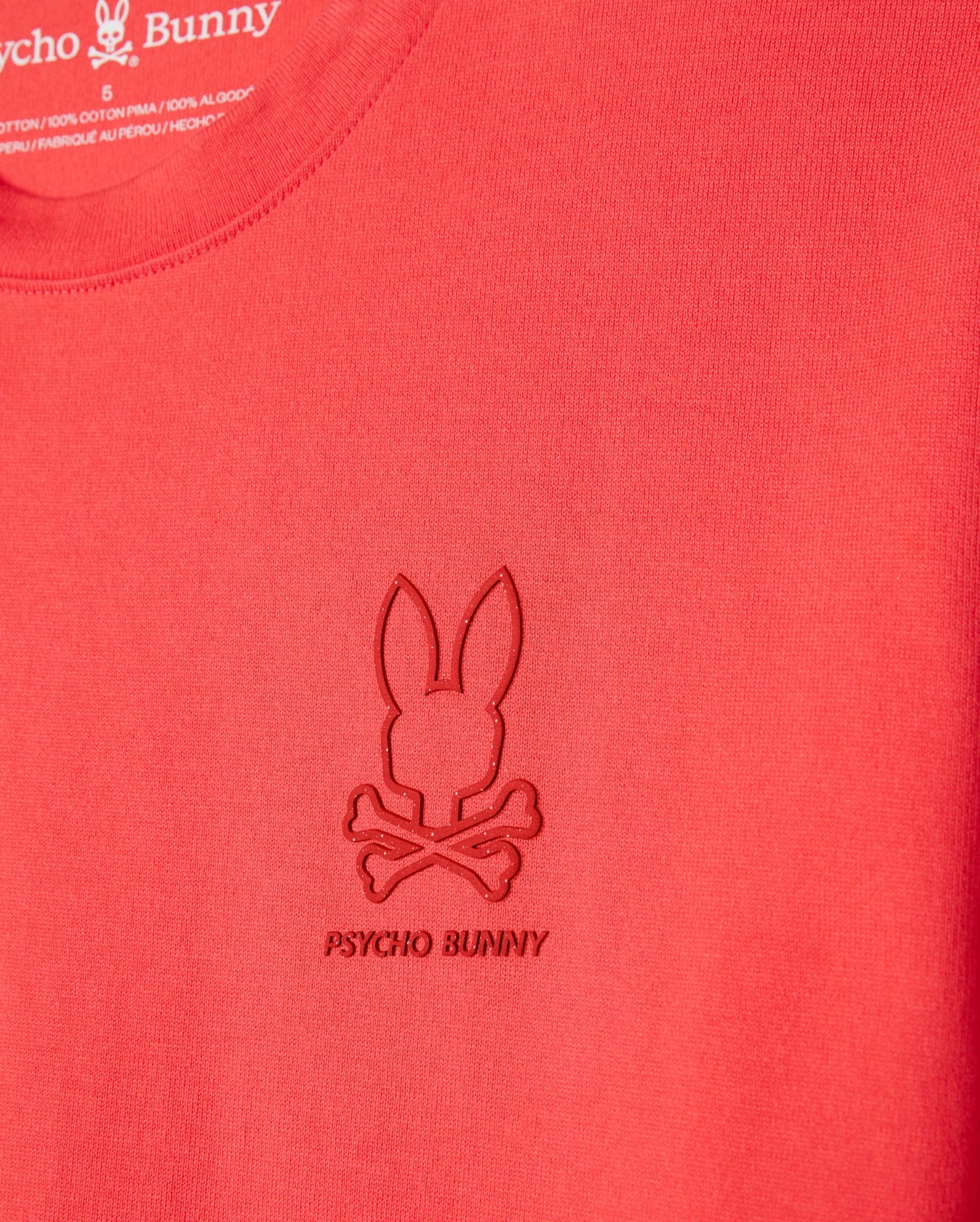 Close-up of a red Psycho Bunny MENS BARRETT RELAXED FIT TEE - B6U518C200 featuring an embossed Bunny graphic with a bunny head and crossbones below it. The text "PSYCHO BUNNY" is stitched under the logo. Made from smooth, heavyweight Pima cotton, the fabric ensures durability while maintaining a relaxed fit.