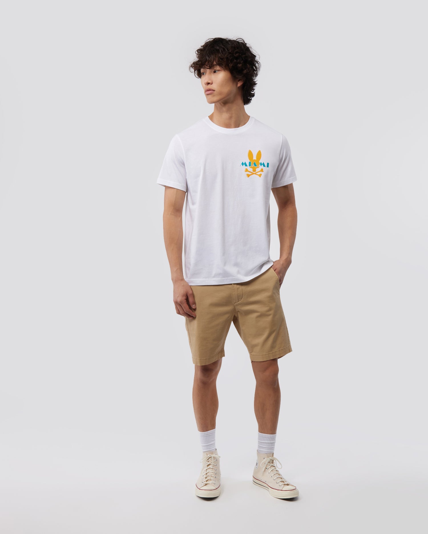 A person with short, curly dark hair is standing against a plain light gray background. They are wearing a Psycho Bunny MENS MIAMI TEE - B6U550W1PC made of soft Pima cotton, beige shorts, white socks, and off-white sneakers. The colorful graphic on the chest adds a vibrant Miami flair as they look slightly to the side.