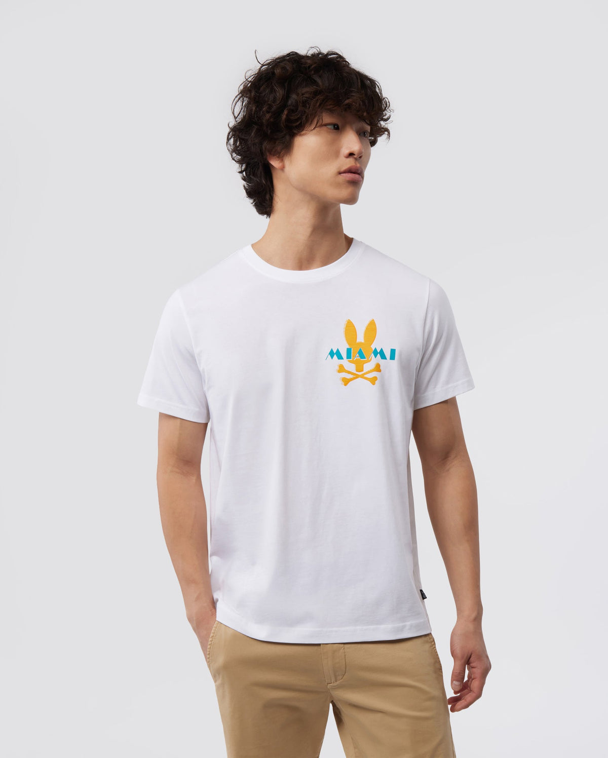 A person with curly hair, wearing a white Psycho Bunny MENS MIAMI TEE - B6U550W1PC crafted from Pima cotton, which features a Miami graphic with a cartoon rabbit and yellow sun, stands against a plain backdrop. They have their right hand in their beige pants pocket and their head turned slightly to the side.