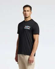 A man is standing against a plain background, wearing the Psycho Bunny MENS CHICAGO TEE - B6U551W1PC in black. This Pima cotton shirt features a small logo on the chest with a cartoonish bird and star design. He has short hair and looks directly at the camera, pairing it with light-colored pants.
