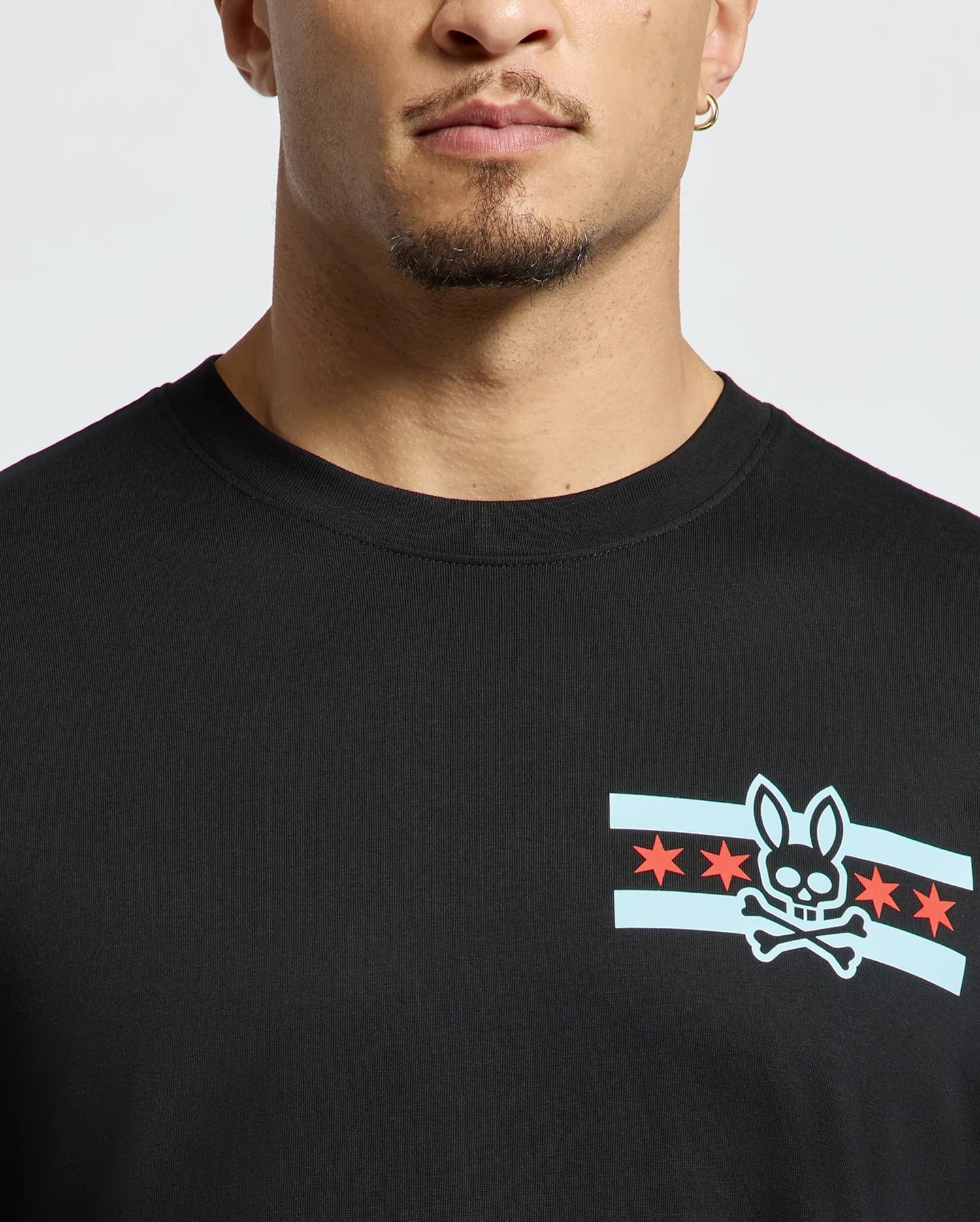 A person wearing a stylish Men's Chicago Tee by Psycho Bunny in black, featuring a graphic design of a bunny skull and crossbones surrounded by blue stripes and red stars. The image highlights the upper portion of this shirt.