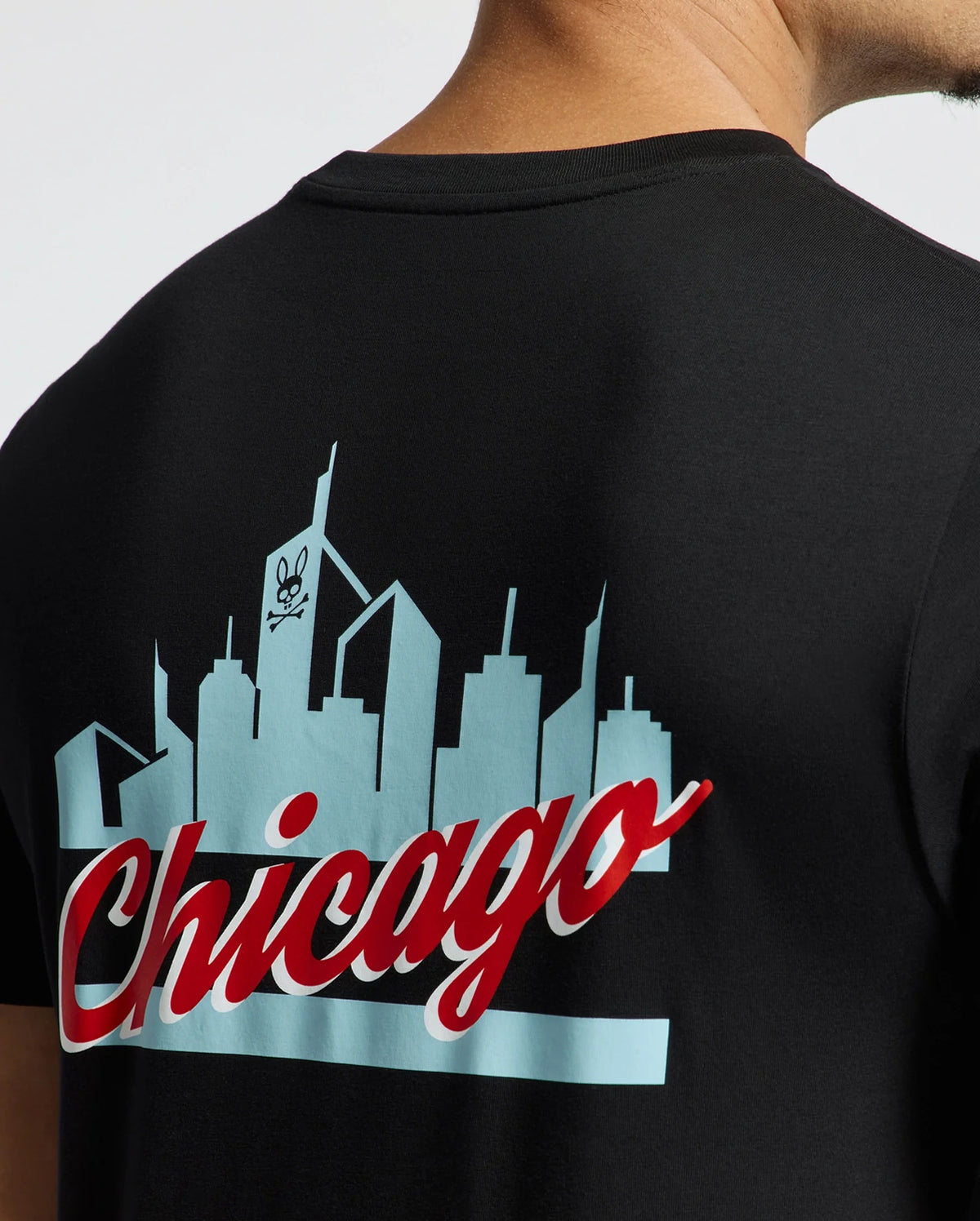 A person is wearing the sleek black MENS CHICAGO TEE - B6U551W1PC from Psycho Bunny, crafted from soft Pima cotton. It features a striking skyline graphic with a skull logo and "Chicago" boldly printed in red and white. The view shows part of the neck and shoulder from behind.