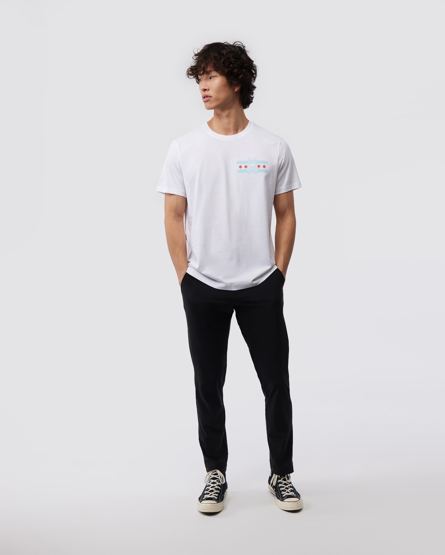 Against a minimal backdrop, an individual with curly hair is styled in a Psycho Bunny's white MENS CHICAGO CITY TEE - B6U551W1PC, featuring a small, vibrant Bunny logo graphic. The outfit is enhanced with black pants and black sneakers with contrasting white soles, made from soft Pima cotton jersey. The person nonchalantly has their hands in their pockets.