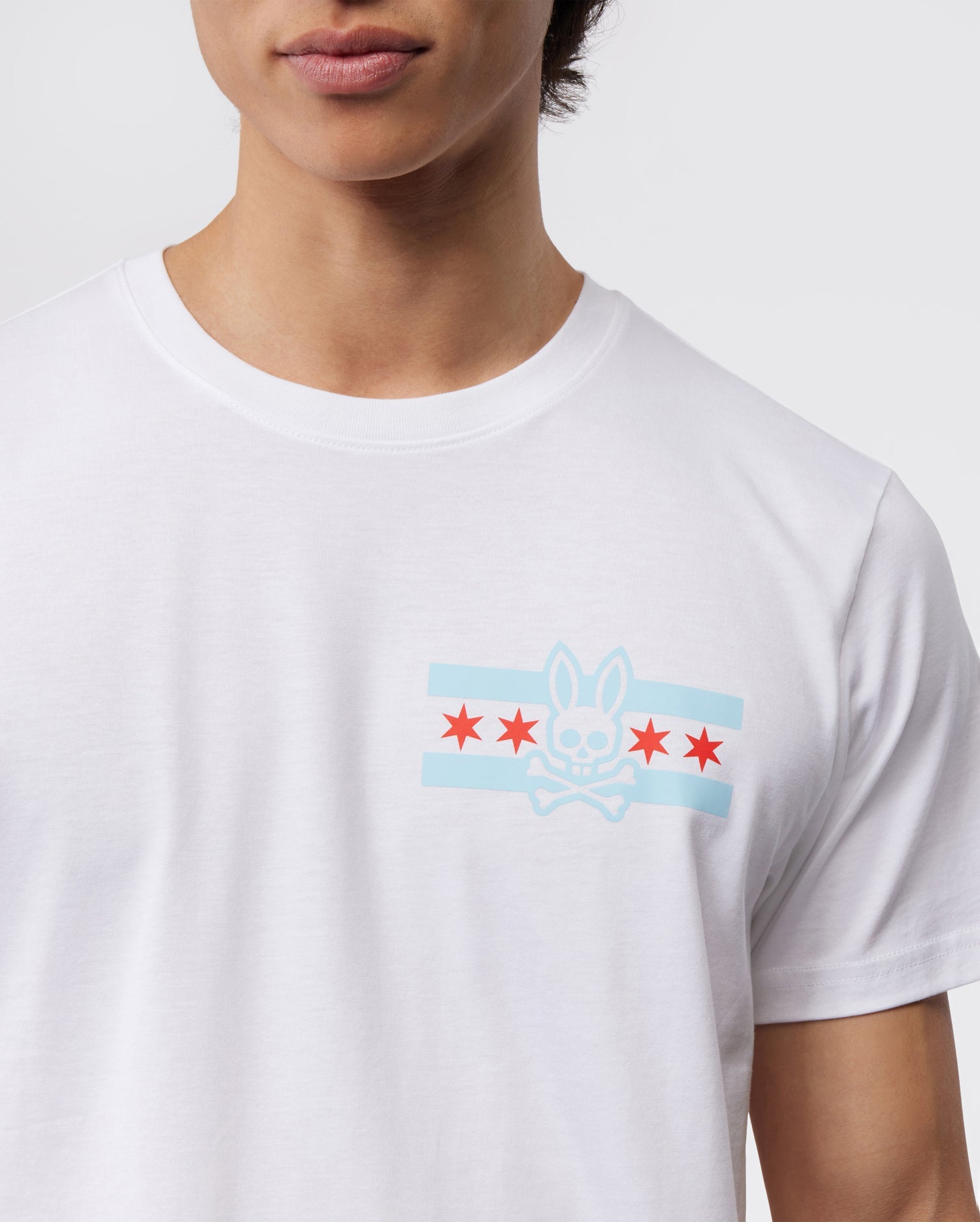 A person wears the Psycho Bunny Men's Chicago City Tee - B6U551W1PC, crafted from soft Pima cotton jersey and featuring a bunny logo graphic. The design includes a stylized bunny skull and crossbones, two light blue horizontal stripes, and five red stars on the chest against a plain background.