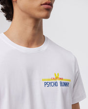 A close-up image showcases a person wearing the MENS CALIFORNIA TEE - B6U552W1PC by Psycho Bunny. This white Pima cotton jersey T-shirt features the words "California Psycho Bunny" printed in blue, red, and yellow with a signature bunny logo above the text. The photo highlights the upper part of this breathable tee and the lower face of the wearer.