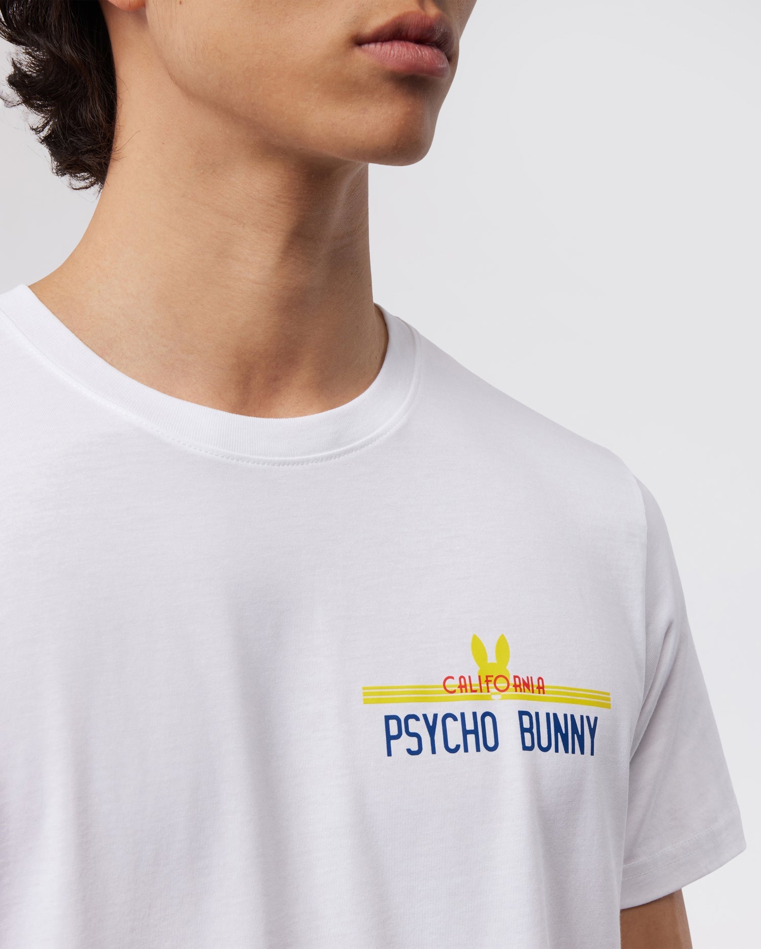 A close-up image showcases a person wearing the MENS CALIFORNIA TEE - B6U552W1PC by Psycho Bunny. This white Pima cotton jersey T-shirt features the words "California Psycho Bunny" printed in blue, red, and yellow with a signature bunny logo above the text. The photo highlights the upper part of this breathable tee and the lower face of the wearer.