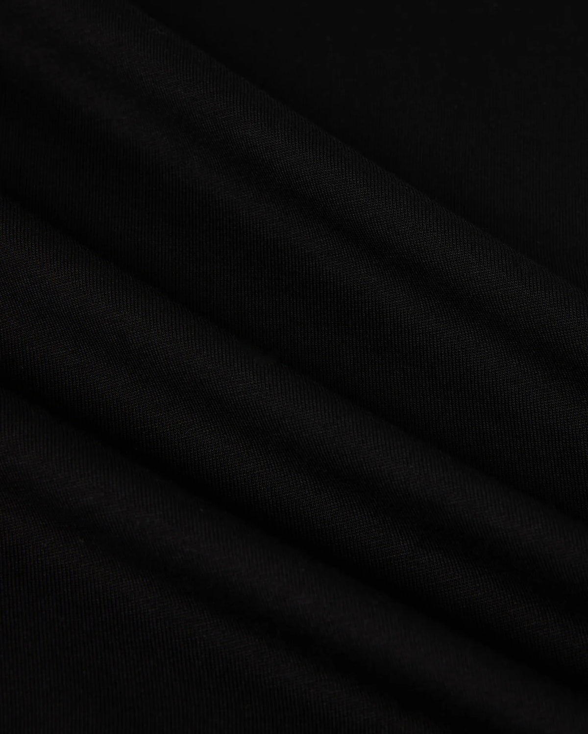 A close-up image of the MENS NEW YORK TEE - B6U554W1PC by Psycho Bunny showcases its smooth, black Pima cotton fabric with soft, diagonal folds. The material has a slightly matte texture and appears to be lightweight. The folds create subtle shadows and highlights, adding depth to the otherwise uniform color. A small Bunny logo is visible in the corner.
