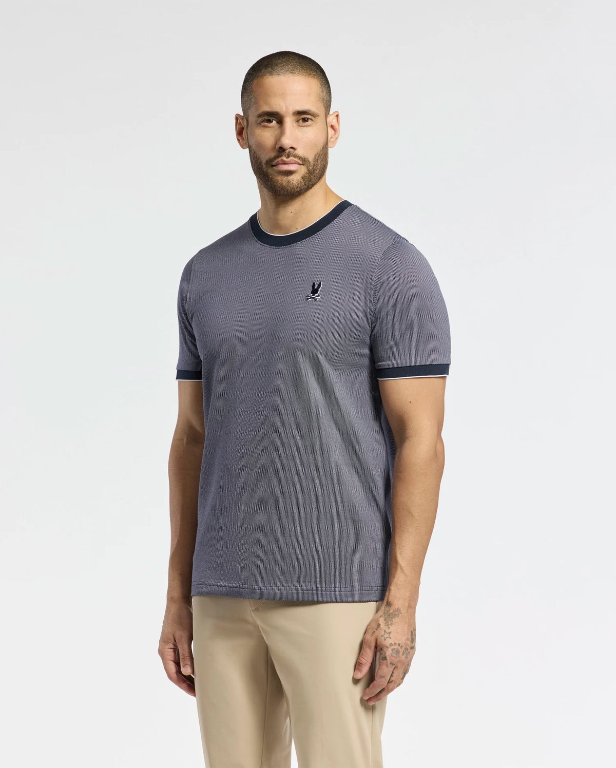 A man with a beard stands against a plain background, wearing the Psycho Bunny MENS MACKAY JACQUARD FASHION TEE, showcasing an embroidered chest Bunny and black trim, paired with beige pants. The premium fabric drapes well, while a subtle tattoo is visible on his left hand as his arms rest casually.