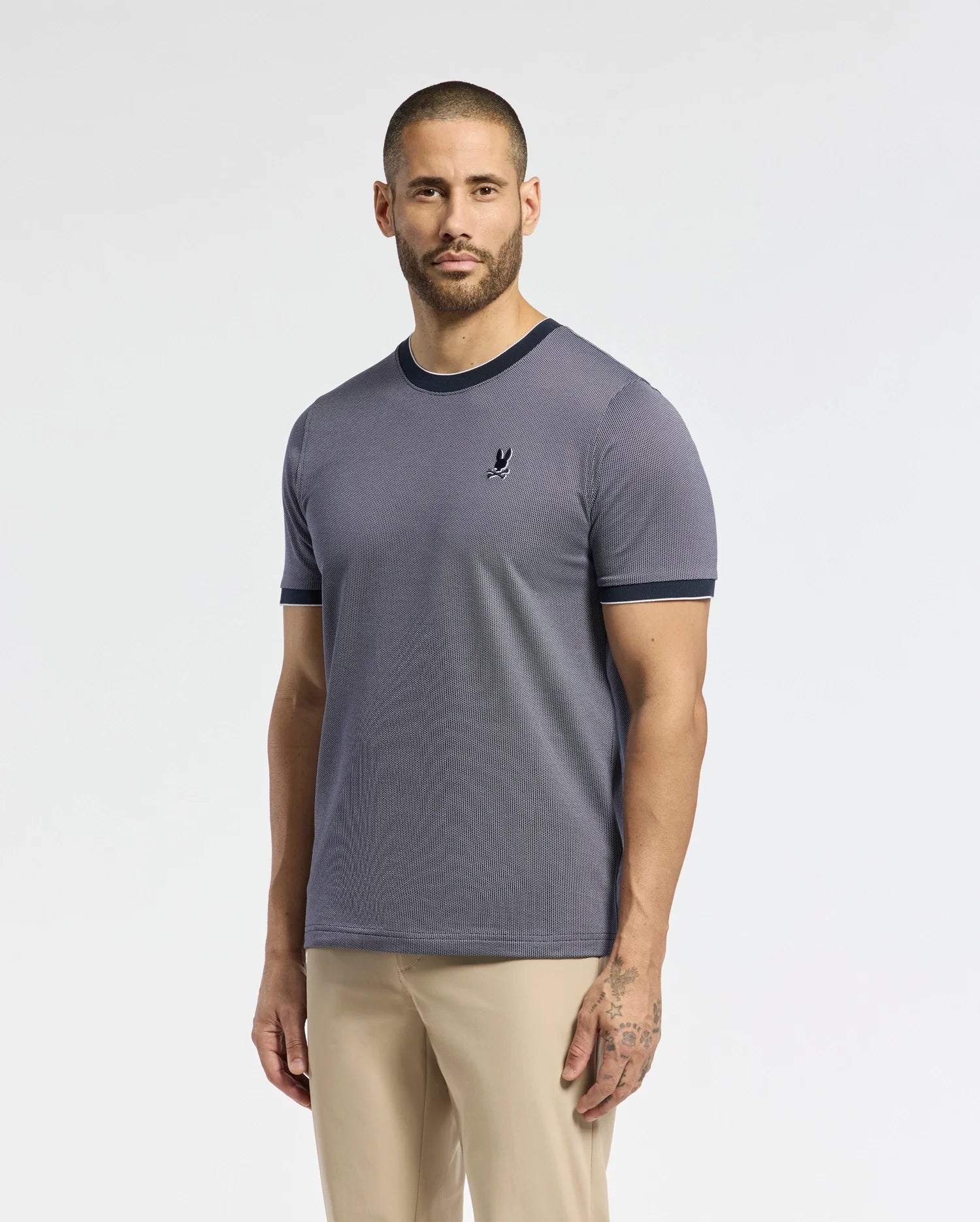 A man with a beard stands against a plain background, wearing the Psycho Bunny MENS MACKAY JACQUARD FASHION TEE, showcasing an embroidered chest Bunny and black trim, paired with beige pants. The premium fabric drapes well, while a subtle tattoo is visible on his left hand as his arms rest casually.