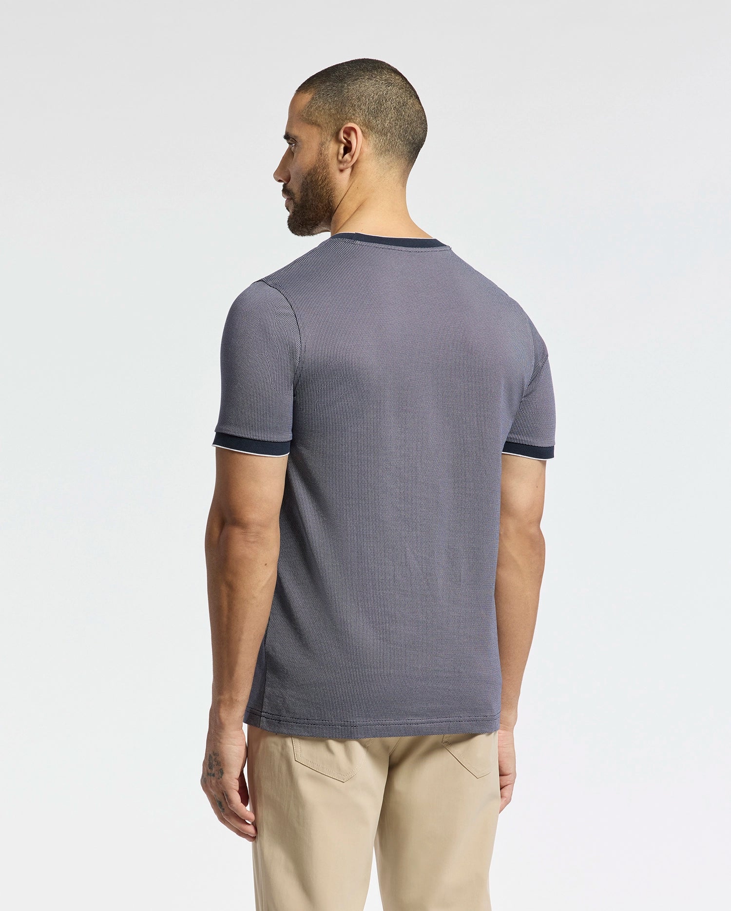 A man with short hair and a beard stands sideways, looking away, in a Psycho Bunny MENS MACKAY JACQUARD FASHION TEE (B6U576F200) made of premium gray Pima cotton with dark trim. Beige pants complement his outfit. His left arm has a small tattoo, and an embroidered chest Bunny adds detail against the plain white background.