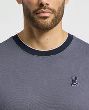 A man wears the Psycho Bunny Men's Mackay Jacquard Fashion Tee, featuring a premium dark gray textured design with a black collar. Made from Jacquard-knit Pima cotton, it showcases an embroidered chest Bunny logo with crossed bones on the left against a white background.