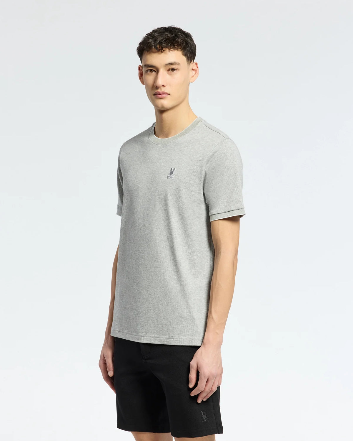A person in a gray MENS MACKAY JACQUARD FASHION TEE by Psycho Bunny, featuring an embroidered chest Bunny, with black shorts stands against a plain white background. The individual has short curly hair and a neutral expression.