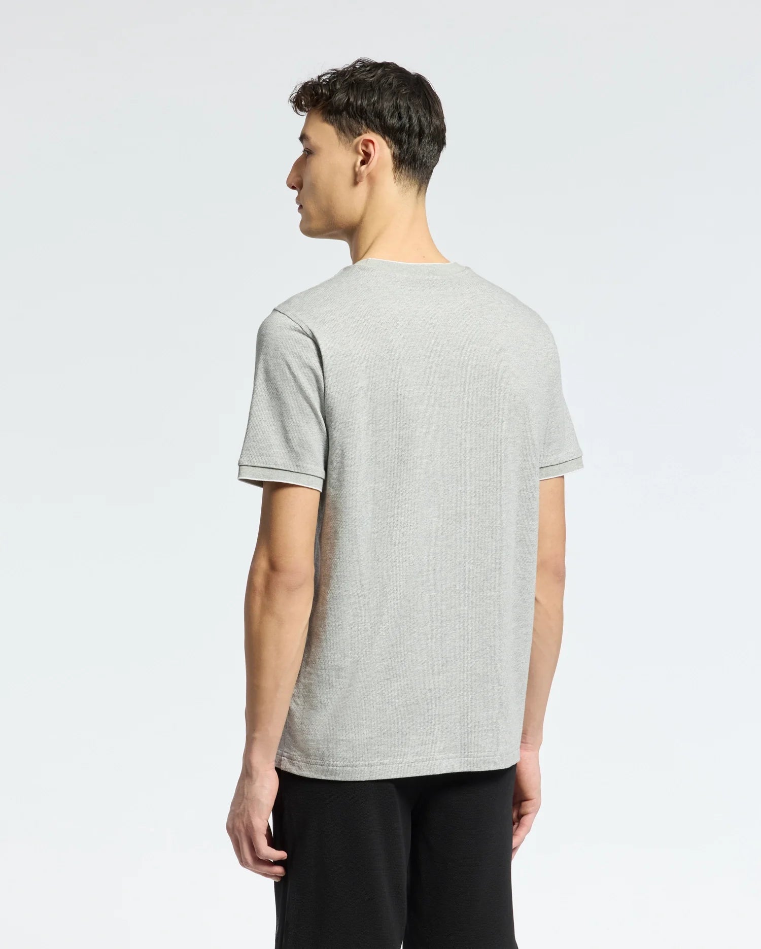 A person with short dark hair stands facing away from the camera against a plain background, wearing a light gray Psycho Bunny MENS MACKAY JACQUARD FASHION TEE - B6U576F200 and black pants.