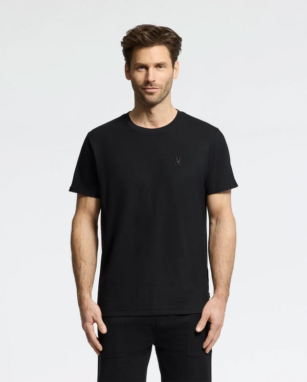 A man with short hair in a Psycho Bunny MENS TAYLOR PIQUE TEE (B6U579F200), wearing matching pants, stands against a white background. The embroidered chest Bunny adds subtle detail as he faces forward with relaxed hands by his sides.