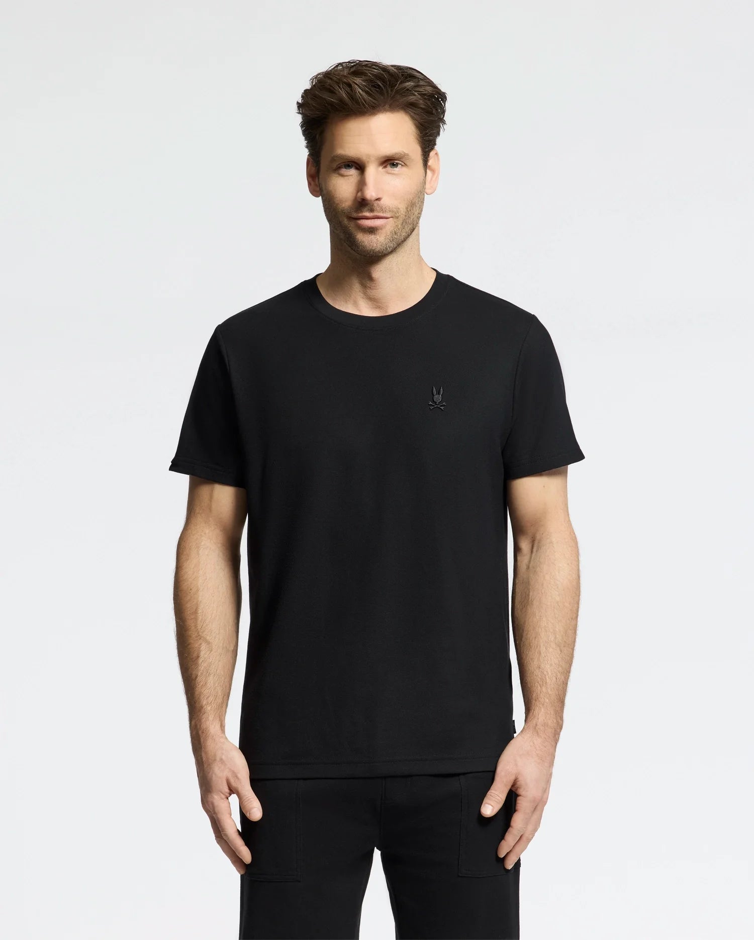 A man with short hair in a Psycho Bunny MENS TAYLOR PIQUE TEE (B6U579F200), wearing matching pants, stands against a white background. The embroidered chest Bunny adds subtle detail as he faces forward with relaxed hands by his sides.