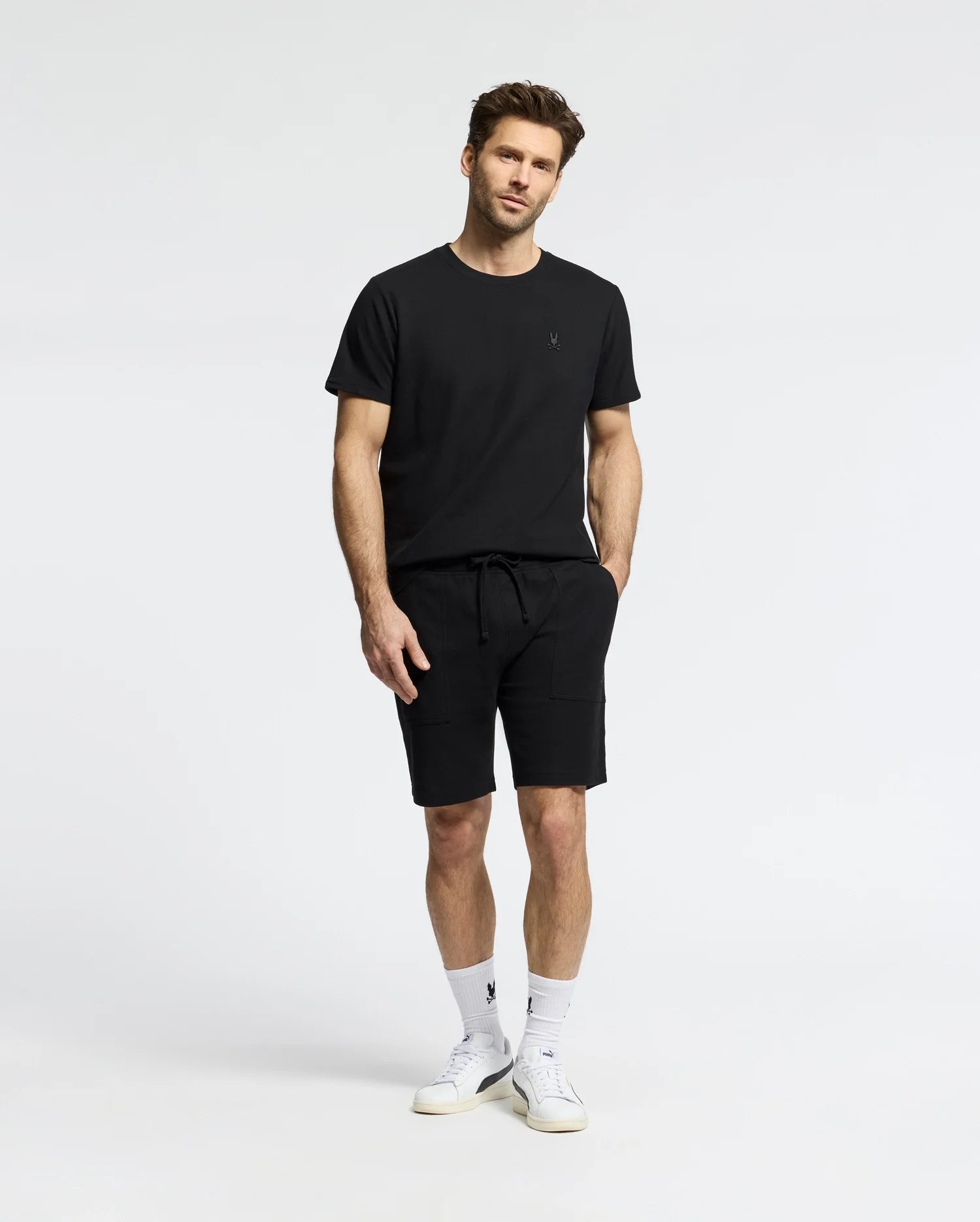 Against a white backdrop, a man wears the MENS TAYLOR PIQUE TEE by Psycho Bunny in black Pima cotton, paired with black shorts. He has his hands in his pockets and complements the look with white socks, sneakers, and a subtle embroidered Bunny on the chest for elegance.