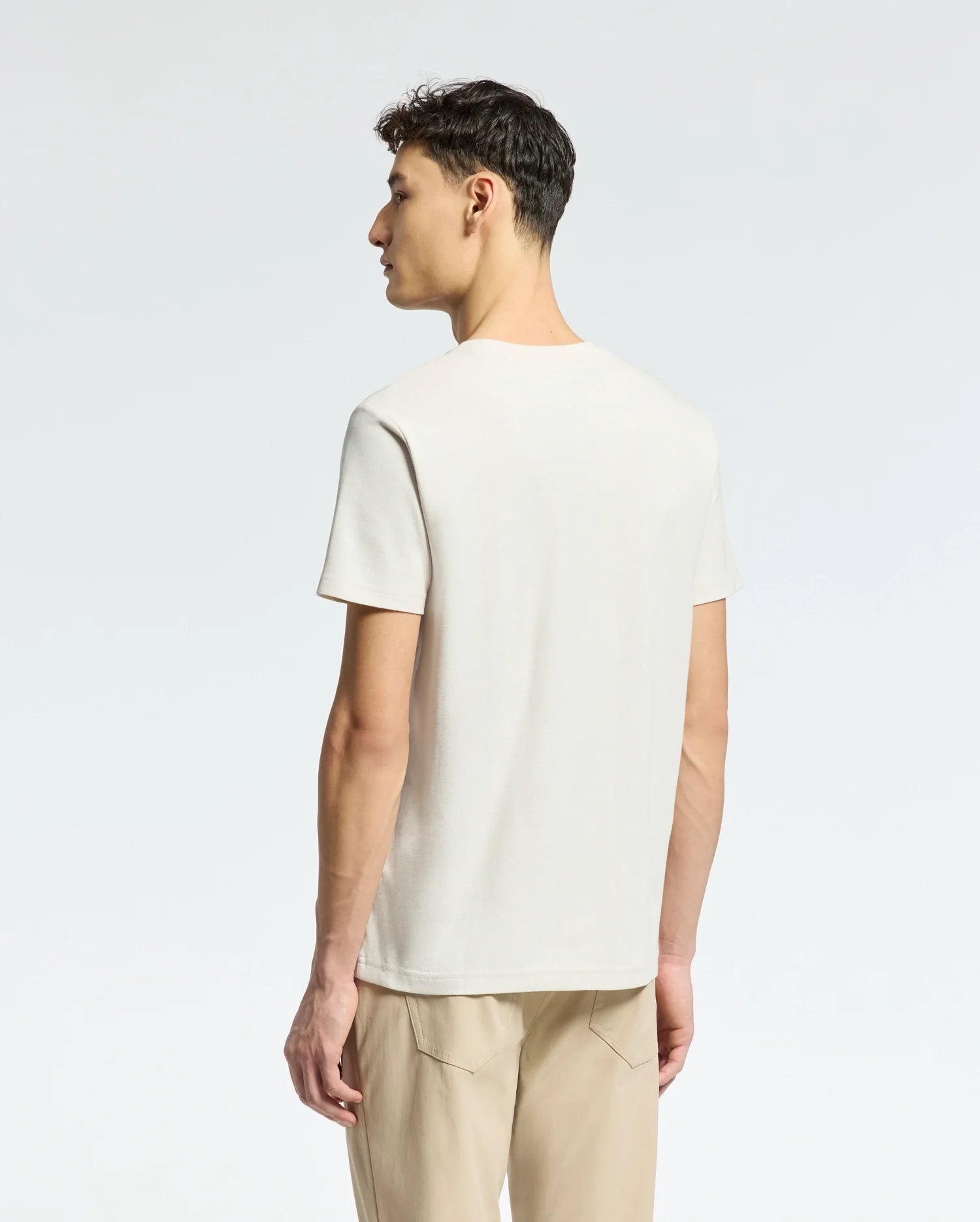 A person stands with their back to the camera, wearing a light beige Psycho Bunny MENS TAYLOR PIQUE TEE, crafted from lightweight Pima cotton, and tan pants. The regular fit tee features a subtle embroidered chest Bunny. The background is a plain, light color.