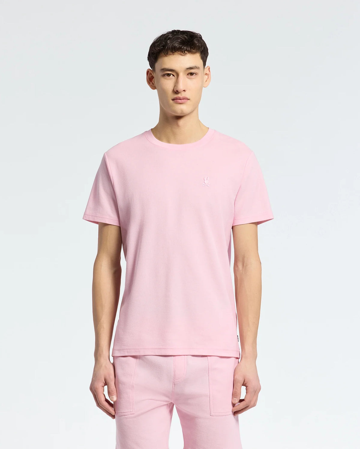 A man in a light pink Psycho Bunny Pima cotton T-shirt and pants stands against a white backdrop. The MENS TAYLOR PIQUE TEE features an embroidered chest Bunny design on the upper left, adding charm.