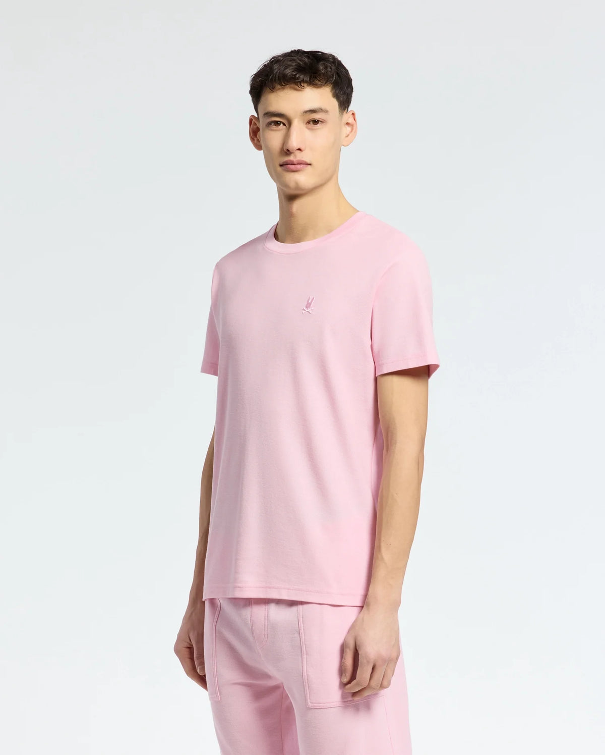 An individual models the Psycho Bunny MENS TAYLOR PIQUE TEE - B6U579F200, paired with matching pants against a plain white backdrop. The light pink piqué T-shirt is lightweight, made from soft Pima cotton, and adorned with an embroidered chest Bunny logo for a playful accent.