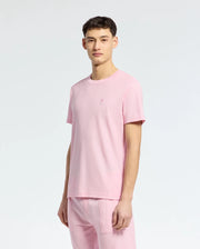 An individual models the Psycho Bunny MENS TAYLOR PIQUE TEE - B6U579F200, paired with matching pants against a plain white backdrop. The light pink piqué T-shirt is lightweight, made from soft Pima cotton, and adorned with an embroidered chest Bunny logo for a playful accent.