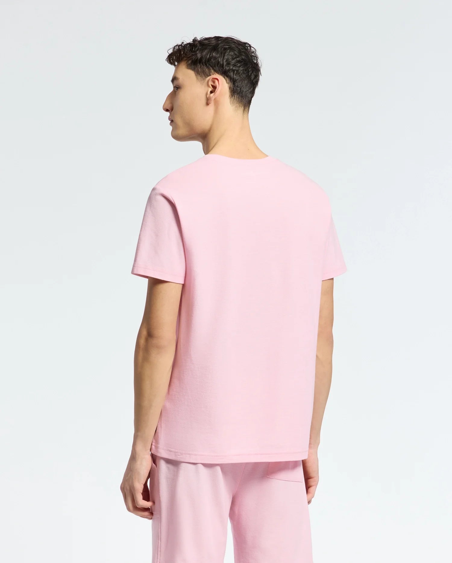 Wearing the MENS TAYLOR PIQUE TEE by Psycho Bunny in pink Pima cotton, a person stands facing away, glancing over their left shoulder. The plain white background sets off the shirt's soft color and subtle embroidered chest Bunny detail.
