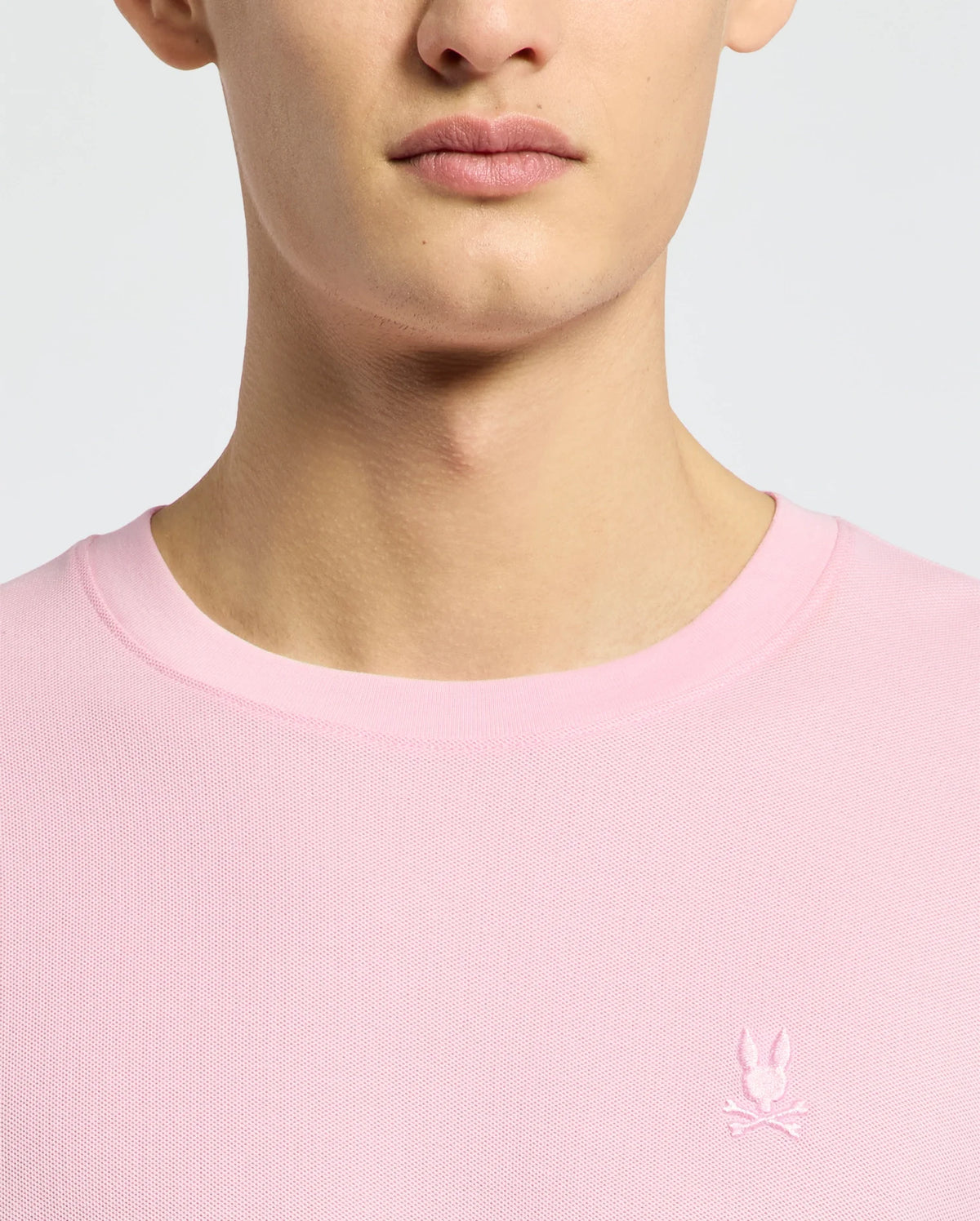Close-up of a person wearing the Psycho Bunny MENS TAYLOR PIQUE TEE - B6U579F200 in light pink, featuring an embroidered white rabbit with crossed bones emblem on the left chest.