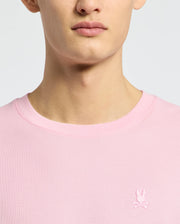 Close-up of a person wearing the Psycho Bunny MENS TAYLOR PIQUE TEE - B6U579F200 in light pink, featuring an embroidered white rabbit with crossed bones emblem on the left chest.