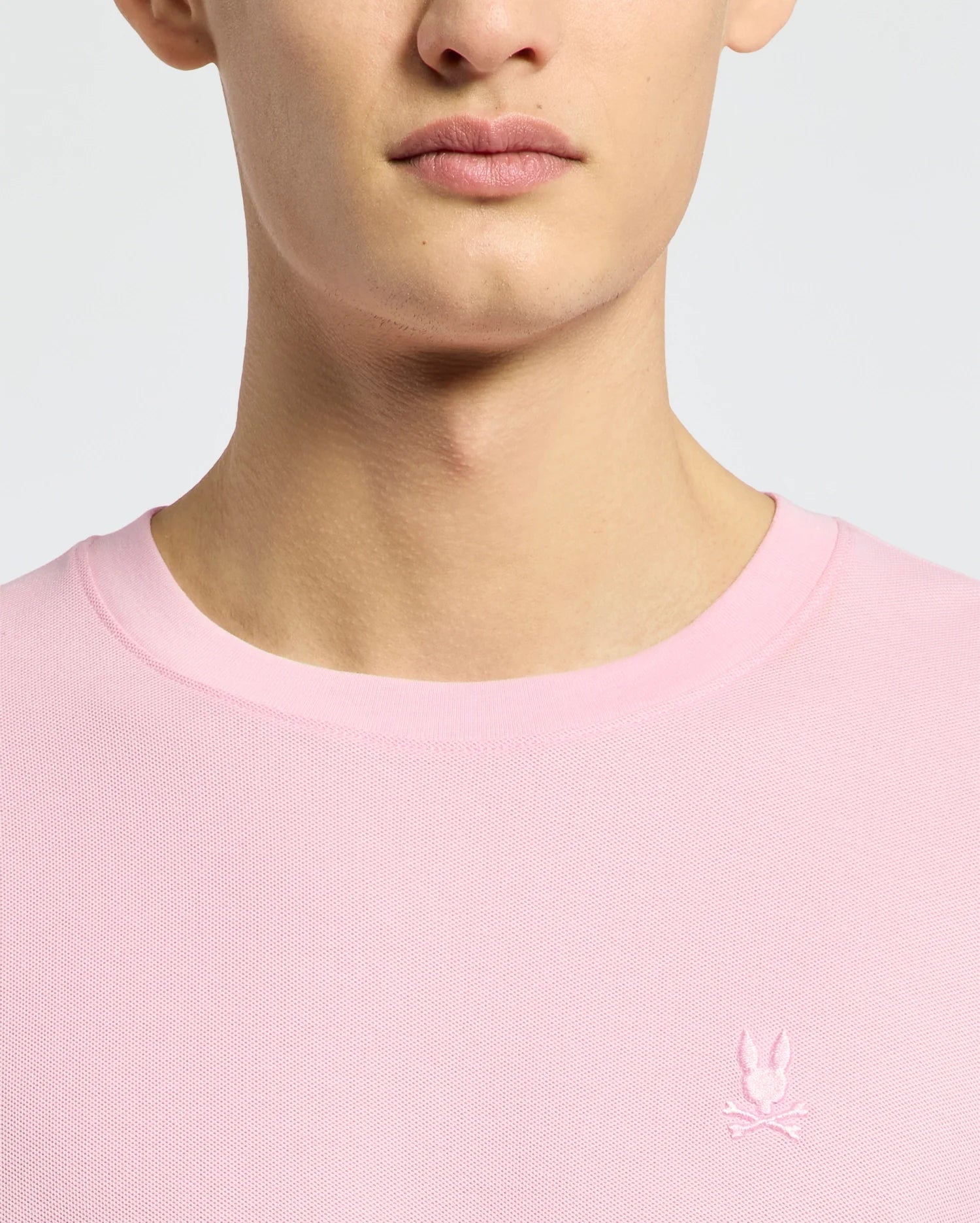 Close-up of a person wearing the Psycho Bunny MENS TAYLOR PIQUE TEE - B6U579F200 in light pink, featuring an embroidered white rabbit with crossed bones emblem on the left chest.
