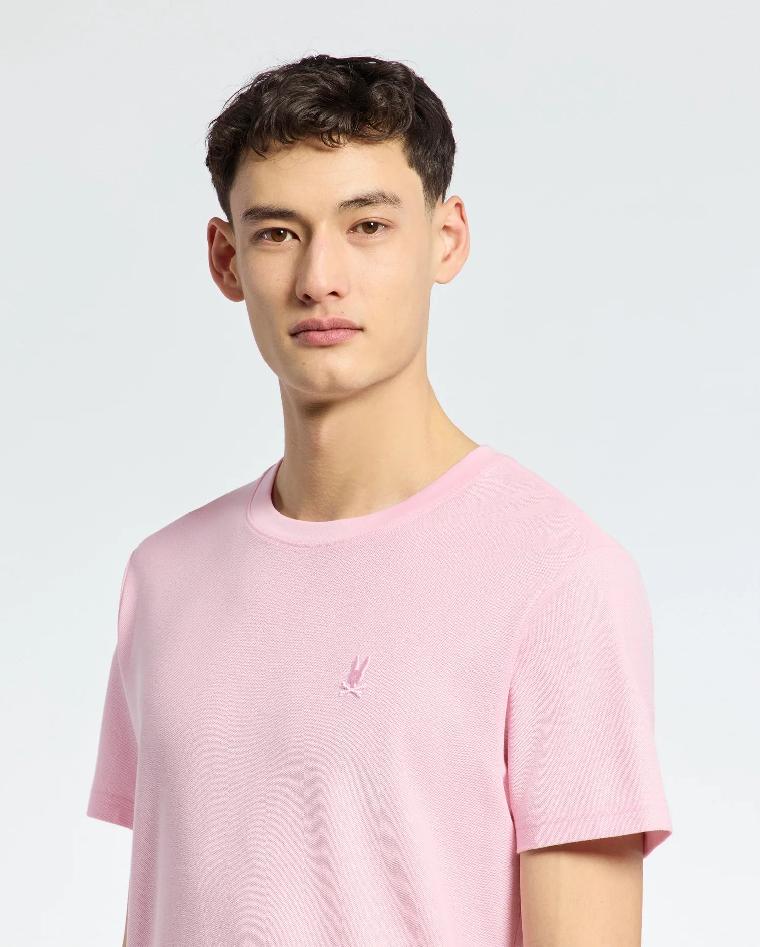 A person in a Psycho Bunny MENS TAYLOR PIQUE TEE - B6U579F200, featuring an embroidered chest Bunny, stands against a light gray backdrop. The individual with short dark hair looks directly at the camera.