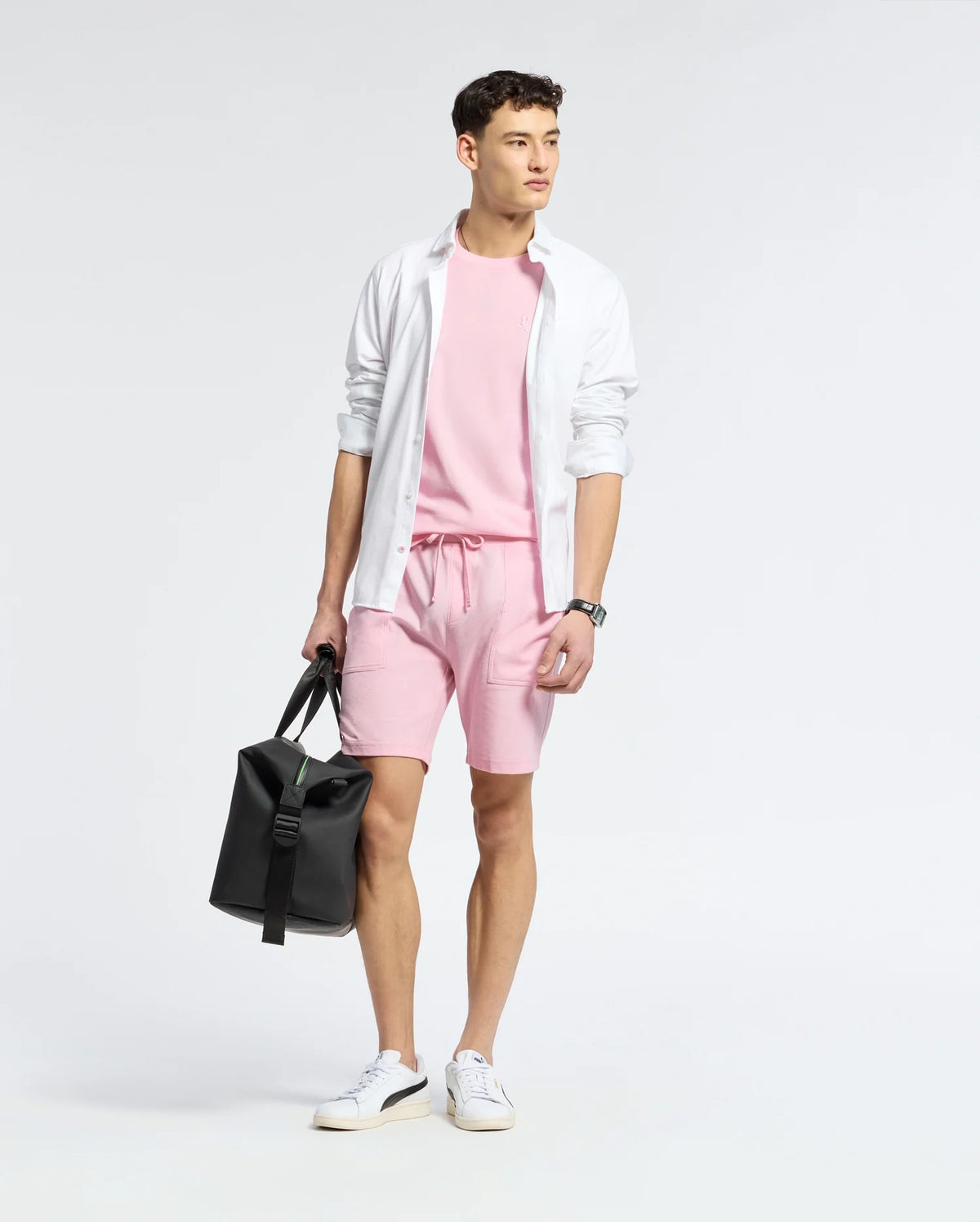 A man stands wearing a light pink TAYLOR PIMA COTTON T-SHIRT by Psycho Bunny, paired with shorts and layered with an open white button-up. An embroidered chest Bunny adds detail. He holds a black bag in his left hand and wears white sneakers against a plain, light backdrop.