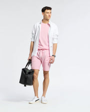 A man stands wearing a light pink TAYLOR PIMA COTTON T-SHIRT by Psycho Bunny, paired with shorts and layered with an open white button-up. An embroidered chest Bunny adds detail. He holds a black bag in his left hand and wears white sneakers against a plain, light backdrop.