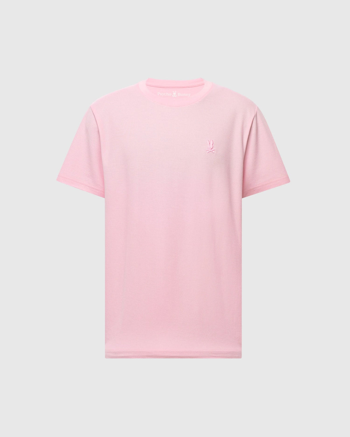 The Psycho Bunny Men's Taylor Piqué Tee, model B6U579F200, is a light pink, short-sleeved shirt made from lightweight piqué fabric. It features an embroidered chest Bunny on the left side and maintains a classic crew neck design for everyday comfort set against a plain gray backdrop.