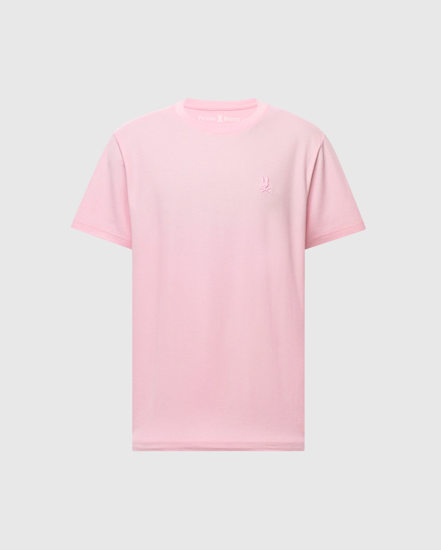 The Psycho Bunny Men's Taylor Piqué Tee, model B6U579F200, is a light pink, short-sleeved shirt made from lightweight piqué fabric. It features an embroidered chest Bunny on the left side and maintains a classic crew neck design for everyday comfort set against a plain gray backdrop.