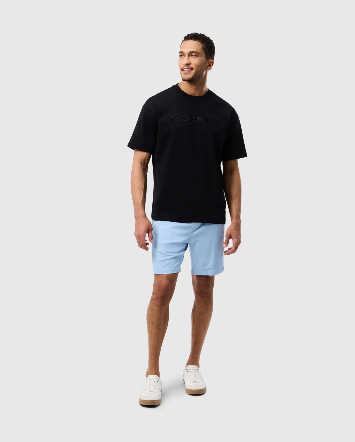 A man wearing a black Psycho Bunny Pima cotton tee with an embroidered "psycho bunny" logo and light blue shorts stands against a white background, looking to the side with a subtle smile. He wears white