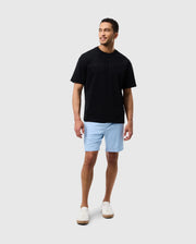 A man wearing a black Psycho Bunny Pima cotton tee with an embroidered "psycho bunny" logo and light blue shorts stands against a white background, looking to the side with a subtle smile. He wears white