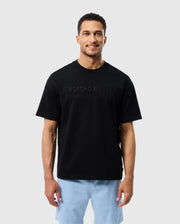 A man smiling at the camera, wearing a black Psycho Bunny Pima cotton tee with an embroidered "psycho bunny" logo and light blue shorts, standing against a white background.