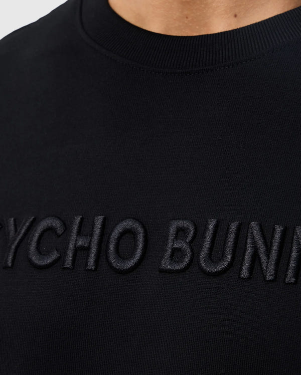 Close-up of a black sweatshirt with the words "MENS WINDCREST HEAVY WEIGHT TEE - B6U593C200" embroidered in black letters on the chest, showing detailed fabric texture and neckline by Psycho Bunny.