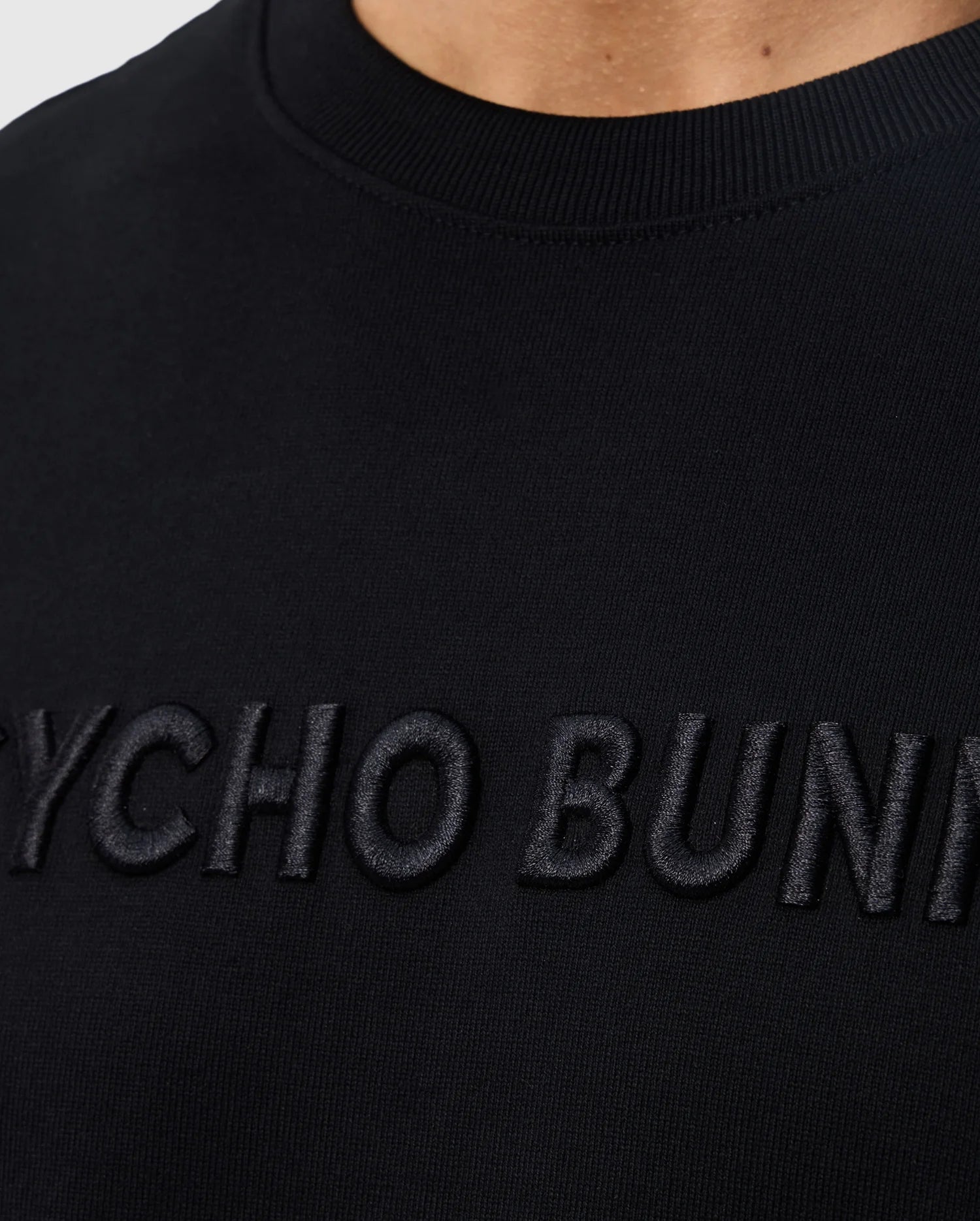 Close-up of a black sweatshirt with the words "MENS WINDCREST HEAVY WEIGHT TEE - B6U593C200" embroidered in black letters on the chest, showing detailed fabric texture and neckline by Psycho Bunny.