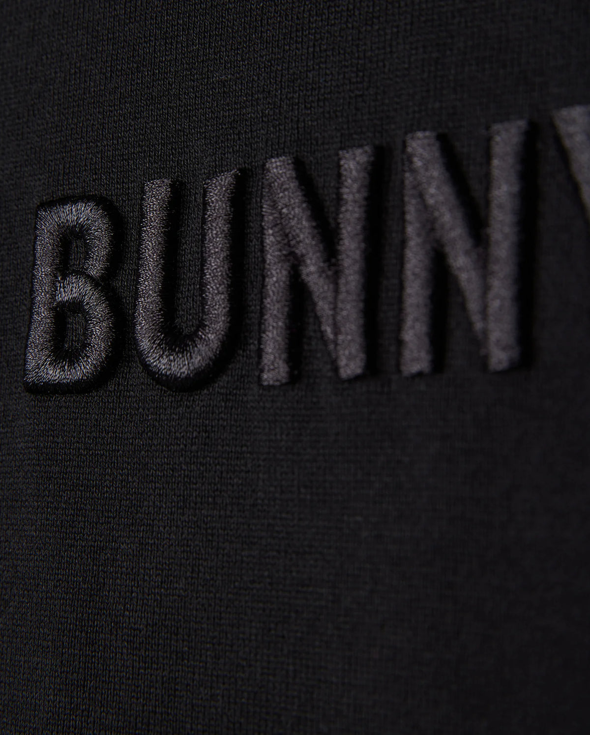 Close-up of the word "Psycho Bunny" embroidered logo in black thread on a black fabric background, with a focus on texture and detail.