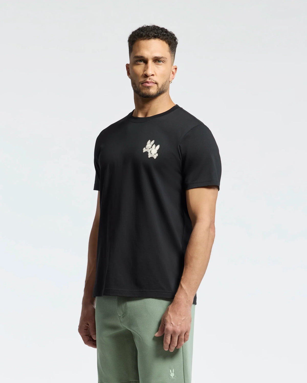 A man stands facing forward, wearing a Psycho Bunny Men's Elliot Front Back Graphic Tee in black, crafted from soft Pima cotton with a small water-based chest print, paired with light green pants. The background is plain and light-colored.