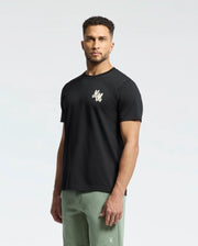 A man stands facing forward, wearing a Psycho Bunny Men's Elliot Front Back Graphic Tee in black, crafted from soft Pima cotton with a small water-based chest print, paired with light green pants. The background is plain and light-colored.