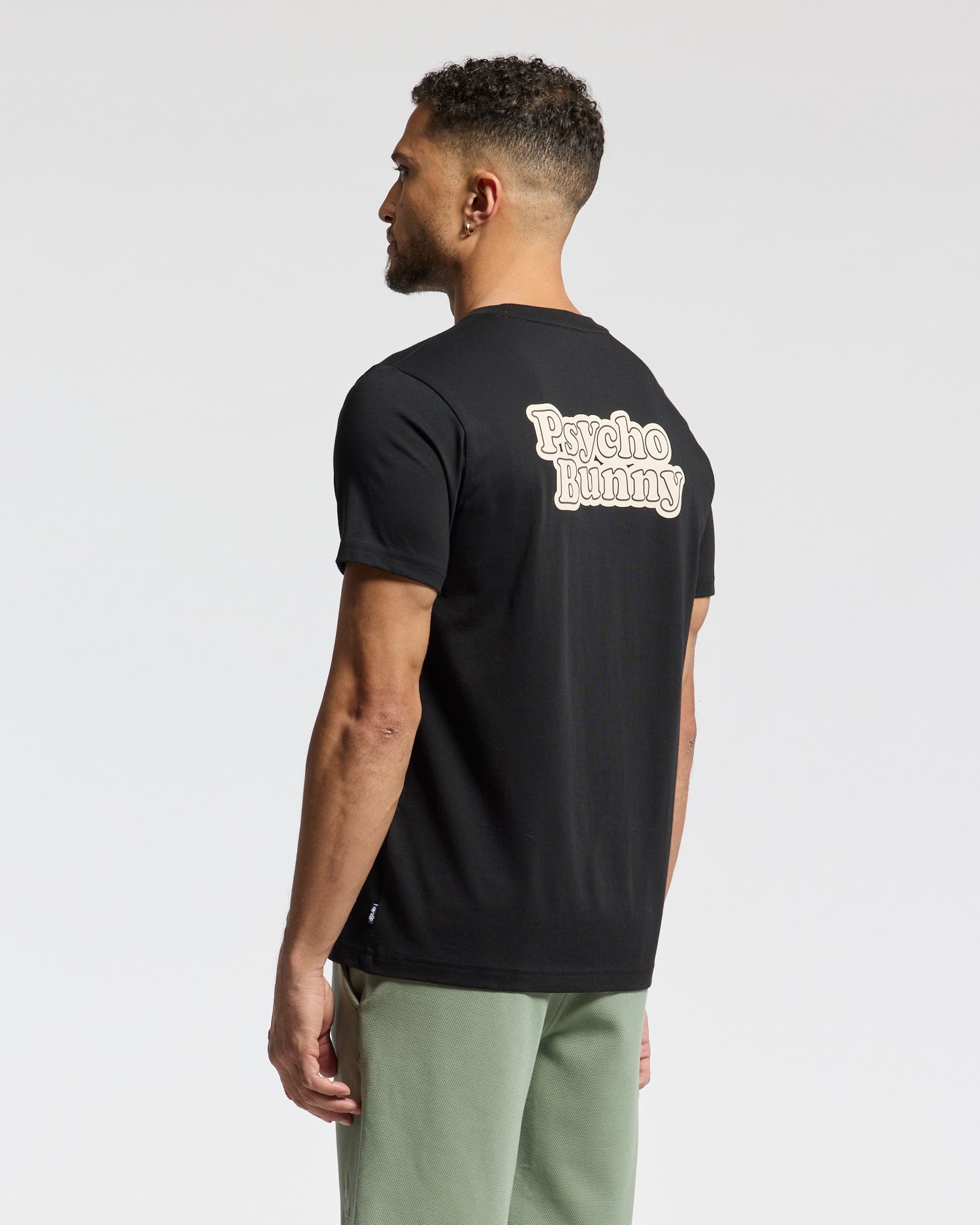 A man with short curly hair, seen from behind, wears the Psycho Bunny MENS ELLIOT FRONT BACK GRAPHIC TEE in black featuring a water-based print. He pairs it with olive pants against a plain white background, showcasing an effortless style.