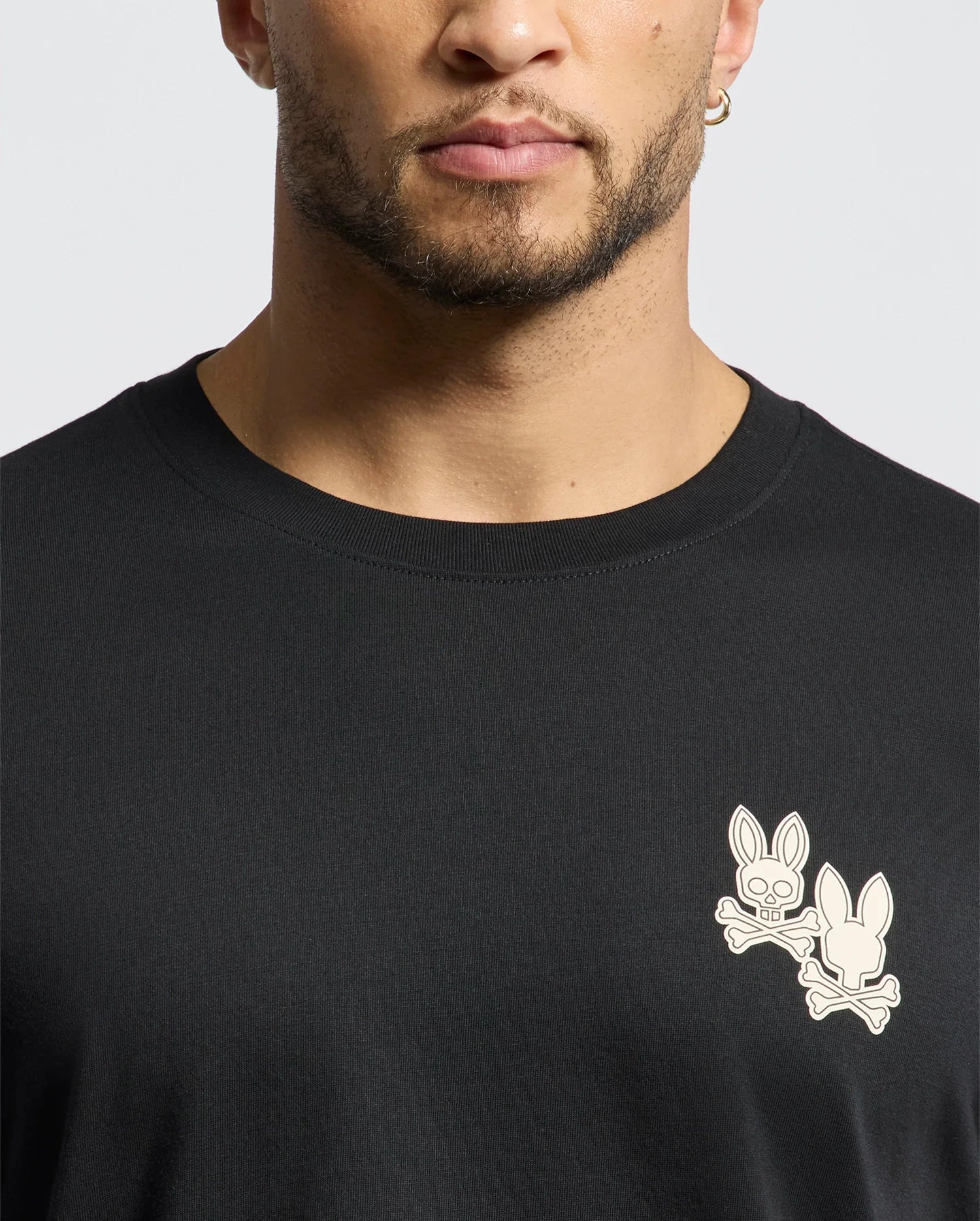 A bearded individual, wearing a hoop earring, sports the Psycho Bunny "Mens Elliot Front Back Graphic Tee - B6U605F200," made from soft Pima cotton with a white bunny and crossbones logo in a water-based chest print, against a plain, light-colored background.