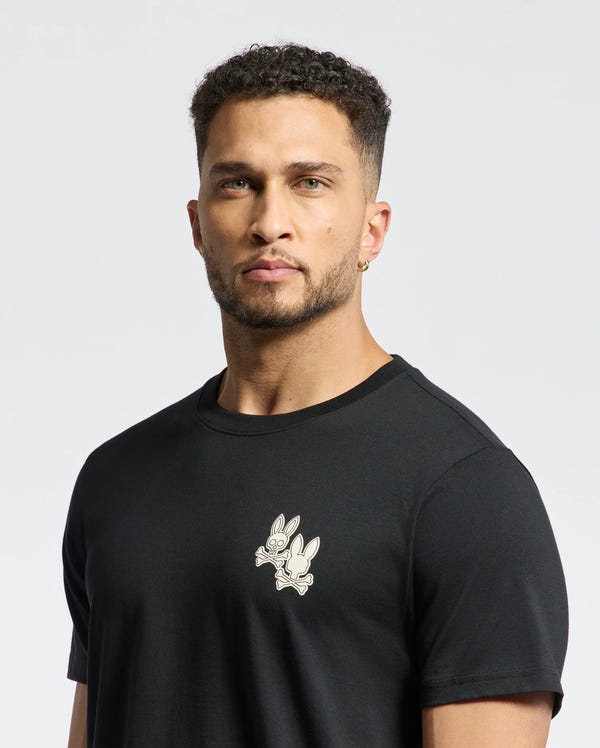 A man with short curly hair and a beard wears the Psycho Bunny MENS ELLIOT FRONT BACK GRAPHIC TEE in black, featuring a small rabbit print. Made from soft Pima cotton, it showcases water-based prints on a white background.