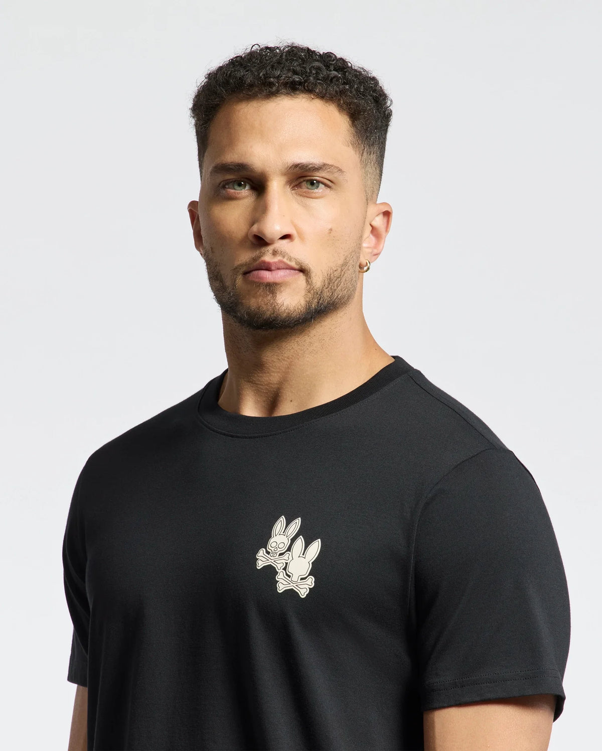 A man with short curly hair and a beard wears the Psycho Bunny MENS ELLIOT FRONT BACK GRAPHIC TEE in black, featuring a small rabbit print. Made from soft Pima cotton, it showcases water-based prints on a white background.