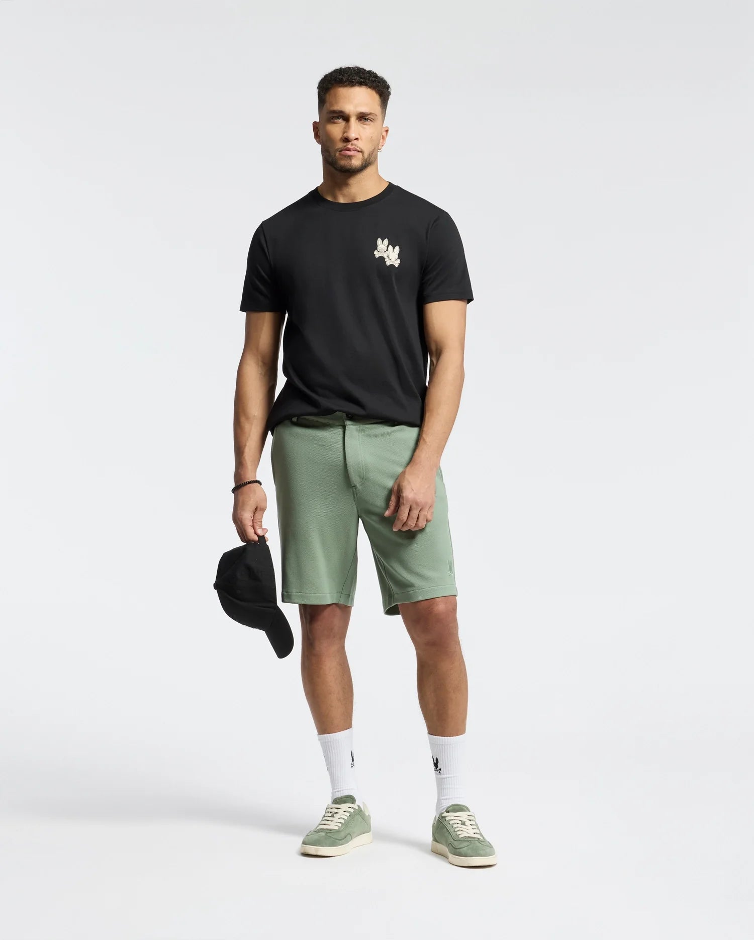 A man in a Psycho Bunny black MENS ELLIOT FRONT BACK GRAPHIC TEE and green shorts stands, holding a black cap in his right hand. He pairs the outfit with white socks and green sneakers, set against a plain white background.