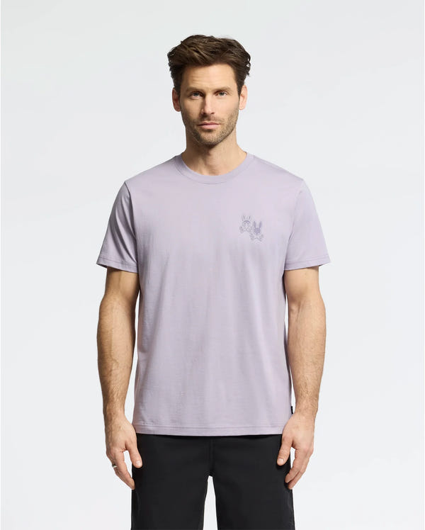A man with short brown hair wears the Psycho Bunny MENS ELLIOT FRONT BACK GRAPHIC TEE in light purple, made of Pima cotton, and black shorts against a plain white background. The regular fit tee features a small graphic print on the upper left side.