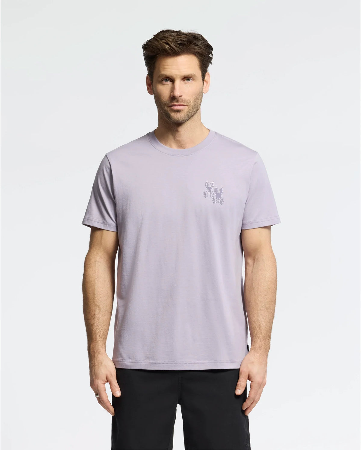 A man with short brown hair wears the Psycho Bunny MENS ELLIOT FRONT BACK GRAPHIC TEE in light purple, made of Pima cotton, and black shorts against a plain white background. The regular fit tee features a small graphic print on the upper left side.