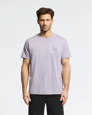 A man with short brown hair wears the Psycho Bunny MENS ELLIOT FRONT BACK GRAPHIC TEE in light purple, made of Pima cotton, and black shorts against a plain white background. The regular fit tee features a small graphic print on the upper left side.