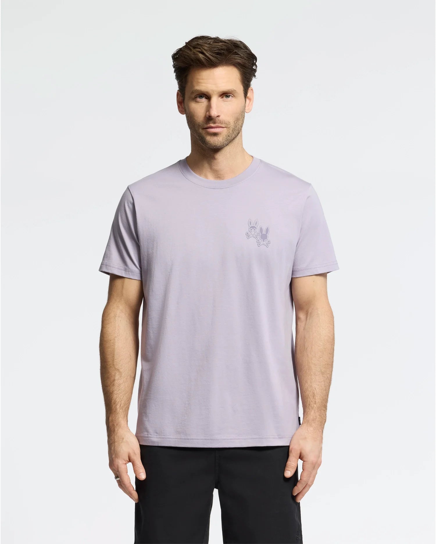 A man with short brown hair wears the Psycho Bunny MENS ELLIOT FRONT BACK GRAPHIC TEE in light purple, made of Pima cotton, and black shorts against a plain white background. The regular fit tee features a small graphic print on the upper left side.